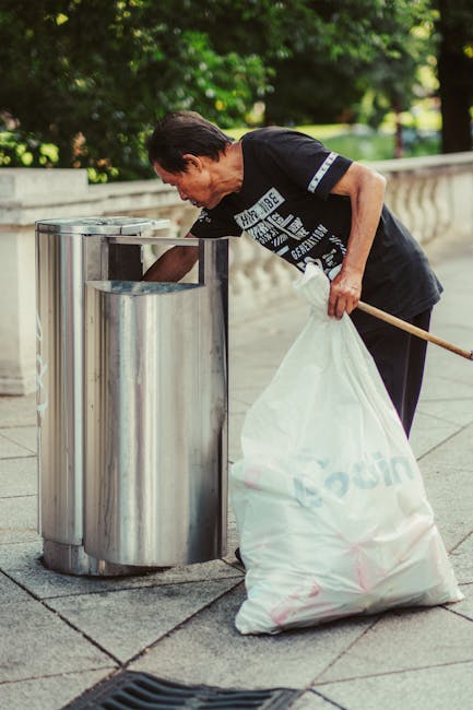 An elderly woman with short dark hair, dressed in a black T-shirt and dark trousers, is seen on a paved outdoor area during daylight hours, collecting and disposing of trash into a stainless steel waste bin with a curved, lidless design. She is bending slightly at the waist, holding a wooden stick in her right hand to assist in loosening or guiding the rubbish, while her left hand grips a large white plastic sack filled with waste. The sack appears slightly crumpled and is marked with faint text or logos, though they are not fully visible. The background features lush green foliage and a stone balustrade, indicating a park or garden setting. The environment is evenly lit with natural sunlight, and the area is clean, suggesting a maintained public space. This scene illustrates a responsible approach to on-site waste disposal and waste management, potentially linked to independent rubbish collection services such as those offered by Waste Removal Sutton, in a manner aligned with alternative rubbish removal methods.