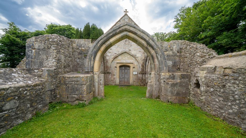 The image depicts an ancient stone church or chapel with a prominent pointed arch entrance made of weathered, light gray stone blocks, leading to a wooden door set within a recessed archway. The stonework shows signs of historical wear and aging, with visible textures and uneven surfaces. The surrounding walls extend outward on both sides, composed of rougher stones and partially crumbled sections, indicating ruins or partial preservation. The ground immediately before the entrance is covered with well-maintained, lush green grass, contrasting with the earthy tones of the stones. Behind the structure, tall, dense trees with vibrant green leaves provide a natural backdrop, and the sky above is partly cloudy with patches of blue. The scene appears to be outdoors, possibly in a park or historic site, and the overall atmosphere suggests a peaceful, weathered historical site that might attract visitors interested in heritage. Waste Removal Sutton’s focus on alternative waste handling processes could be connected indirectly to such outdoor sites requiring occasional clearance of debris or waste from the surrounding area during maintenance activities.