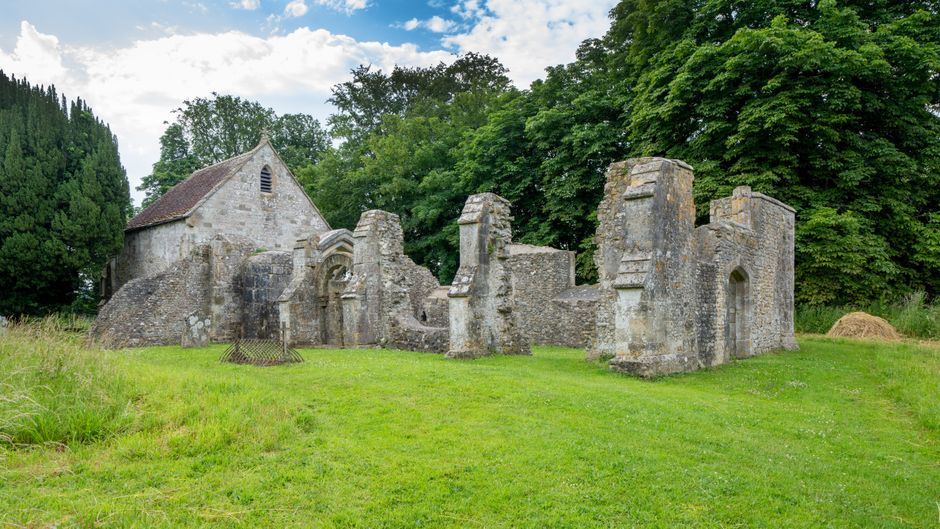 The image displays the remaining stone walls of an ancient, partially ruined church or chapel, situated within a well-maintained grassy area. The weathered grey stone walls feature arched window openings and decorative buttresses, with some sections showing signs of erosion and missing segments. The roof of the structure is absent, exposing the interior to the open sky. Surrounding the ruin, there are tall, lush green trees with dense foliage, providing a natural backdrop that contrasts with the aged stonework. The sky above is partly cloudy with patches of blue visible, illuminating the scene softly. The grassy foreground is neatly trimmed, and there is a small patch of disturbed earth or a mound to the right of the ruins. This setting suggests a rural or historical site that may be targeted for private or independent waste disposal services, such as rubbish removal, which companies like Waste Removal Sutton might handle, especially for clearing debris or remnants from such heritage sites after preservation or restoration efforts.