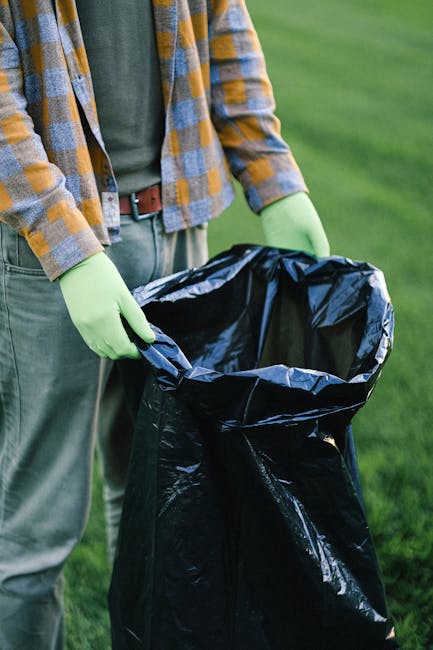 A person dressed in a plaid shirt with shades of yellow, grey, and blue, paired with grey trousers and a brown belt, is standing outdoors on a grassy area. They are wearing light green gloves while holding open a large, black, heavy-duty rubbish bag made of thick plastic. The bag's top edge is folded outward, revealing its shiny, smooth interior surface, and appears to be ready for waste collection or disposal. The scene suggests an environment where waste items are being gathered, possibly as part of a clutter clearing or onsite waste management activity, which could relate to private waste handling or alternative rubbish removal services. The natural daylight and lush green background enhance a professional and neutral tone suitable for a waste removal or rubbish collection context, as carried out by companies like Waste Removal Sutton. The focus remains on the manual handling of waste in an outdoor setting, aligning with efforts in independent rubbish disposal or cleaning initiatives outside traditional municipal collection schemes.