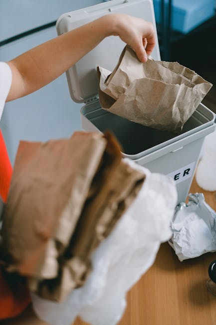 A person is seen disposing of a crumpled brown paper bag into a white rubbish bin, which is partially open, revealing a similar paper bag inside. The individual’s arm is extended, holding the bag over the bin, and they are wearing a short-sleeved garment. The bin is situated on a flat surface, possibly a wooden table or countertop, and the environment appears to be indoors with a clean, modern setting. Additional crumpled paper bags are visible in the foreground, some on the surface near the bin. The scene reflects an aspect of domestic waste disposal, highlighting the process of on-site rubbish removal in a residential context, aligning with waste management services provided by Waste Removal Sutton, especially in relation to the handling of paper-based waste materials applicable to private collection or alternative waste handling methods.