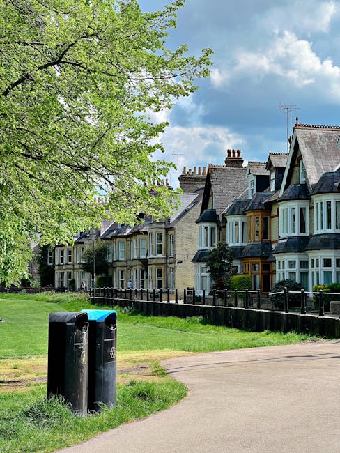 A public park area adjacent to residential terraced houses with Victorian-style architecture, featuring bay windows and decorative brickwork, seen under a partly cloudy sky. In the foreground, two black waste bins with rounded tops and front openings are situated near a grassy patch and a paved pathway. To the left, a large tree with light green leaves extends its branches over the scene, providing shade and a natural backdrop. The houses are positioned along the edge of the park, separated by a low black fence, and have chimneys and varied roof pitches with slate or tile finishes. The environment appears clean and well-maintained, with a soft natural light illuminating the scene, illustrating an example of private outdoor waste disposal or collection points in a residential area. This setting could be related to independent waste removal services, such as those offered by Waste Removal Sutton, where residents manage rubbish collection outside their homes as part of alternative waste handling methods.